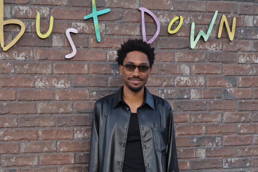 A smiling man in sunglasses, a black jacket and green pants stands in front of a brick wall