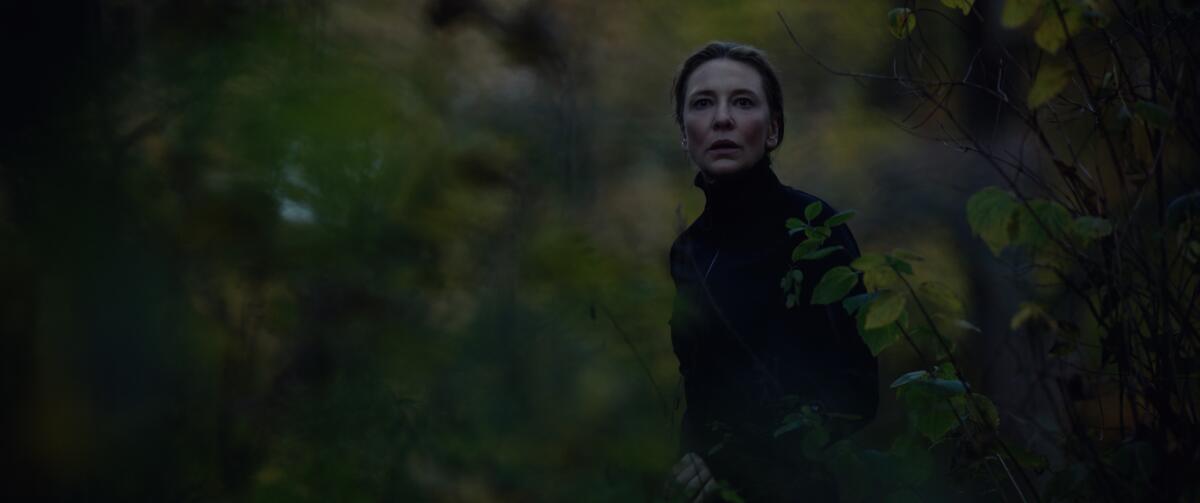 A woman outdoors, wearing a black turtleneck, looks forward with a serious expression.