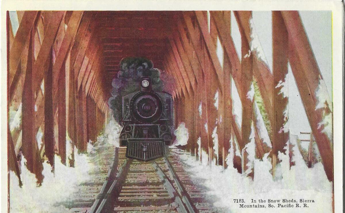 A train barrels toward the viewer, under a structure that prevents snow from accumulating on the tracks