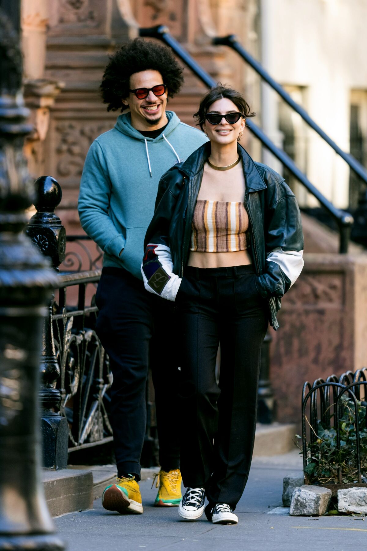 Eric André in glasses and a blue hoodie smiling while walking behind Emily Ratajkowski in a tube top and a big leather jacket
