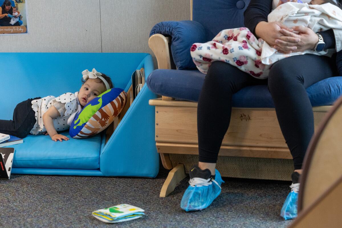 An infant sits on a blue couch next to a woman holding another infant. 