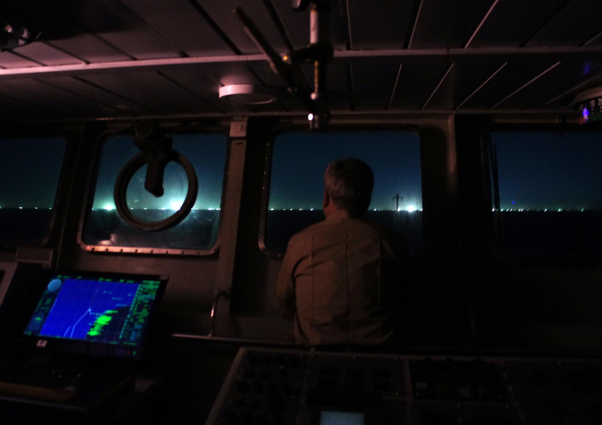 A person stands near the controls of a boat looking out at bright lights in the distance