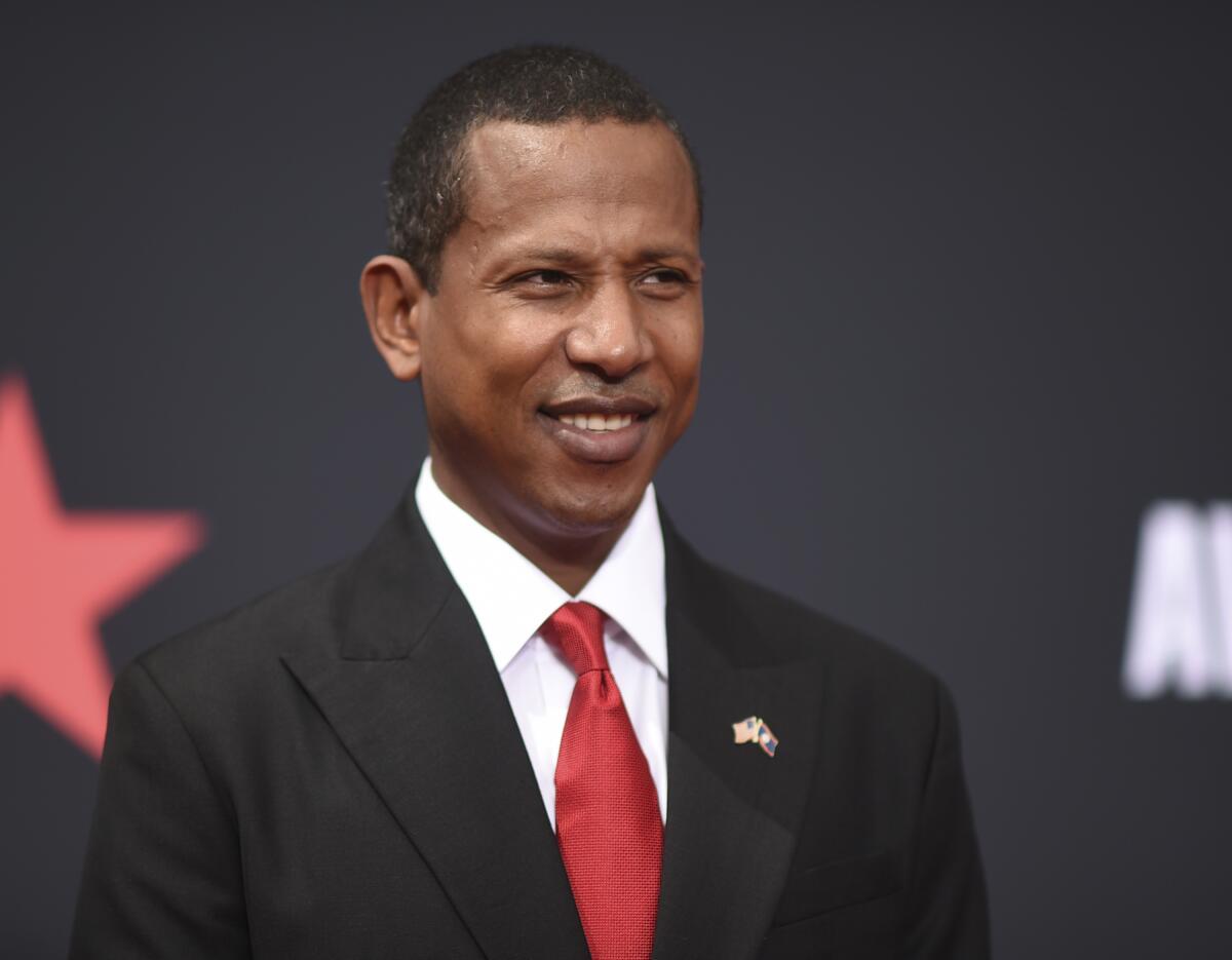 Shyne in a black suit and a red tie smiling and posing against a black background