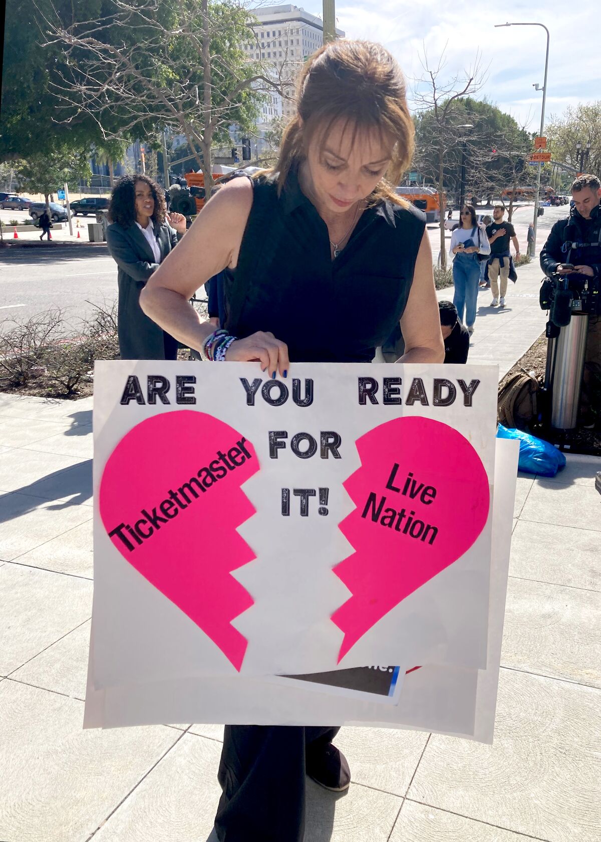 A woman stands outdoors holding a sign with a broken heart that reads, "Are You Ready For It Ticketmaster ... Live Nation"