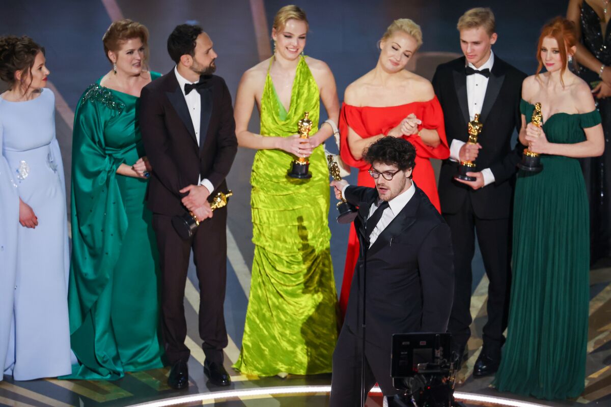 A man wearing formalwear points his Oscar statuette toward other people holding their own statuettes onstage.