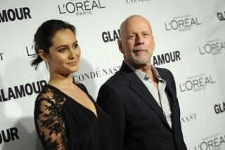 Emma Heming Willis and Bruce Willis pose in formal attire in front of a white backdrop.