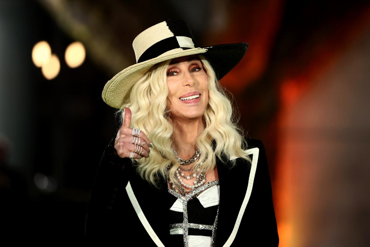 Cher in a blond wig and a black-and-white outfit gives a thumbs-up