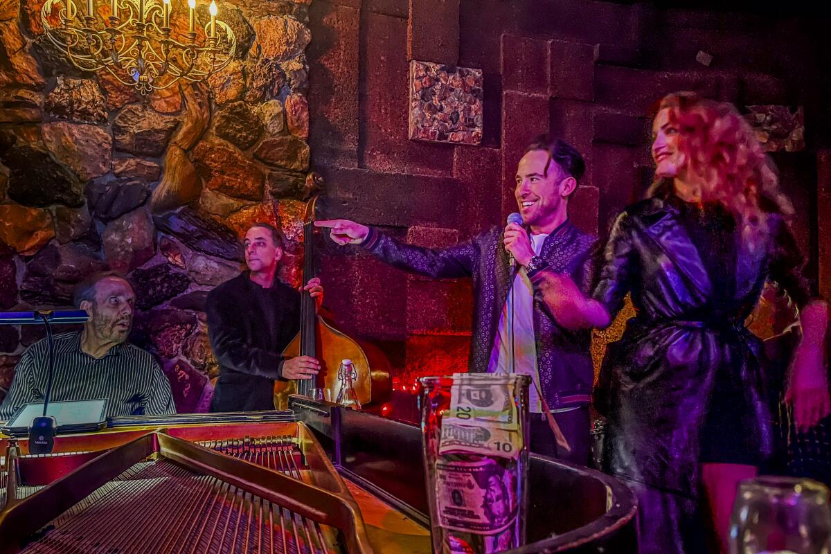 A piano player, a bass player and a male and a female singer around the piano in a bar
