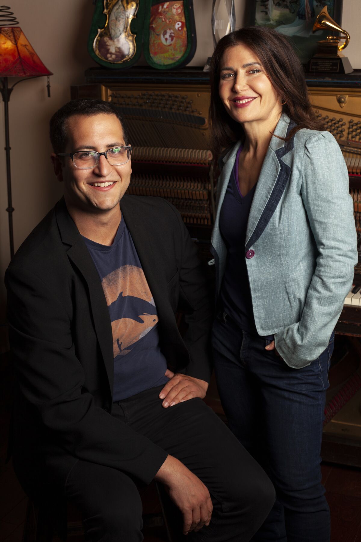 A smiling, bespectacled man in a black blazer & T-shirt sits & a dark-haired, blue-jacketed, smiling woman stands by a piano.