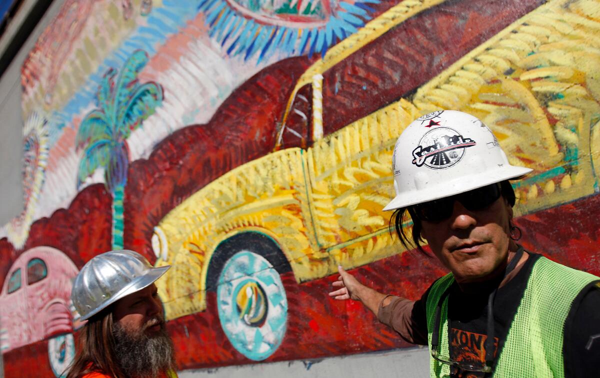 A man in a hard had and safety vest gestures at a mural that shows cars on a roadway.