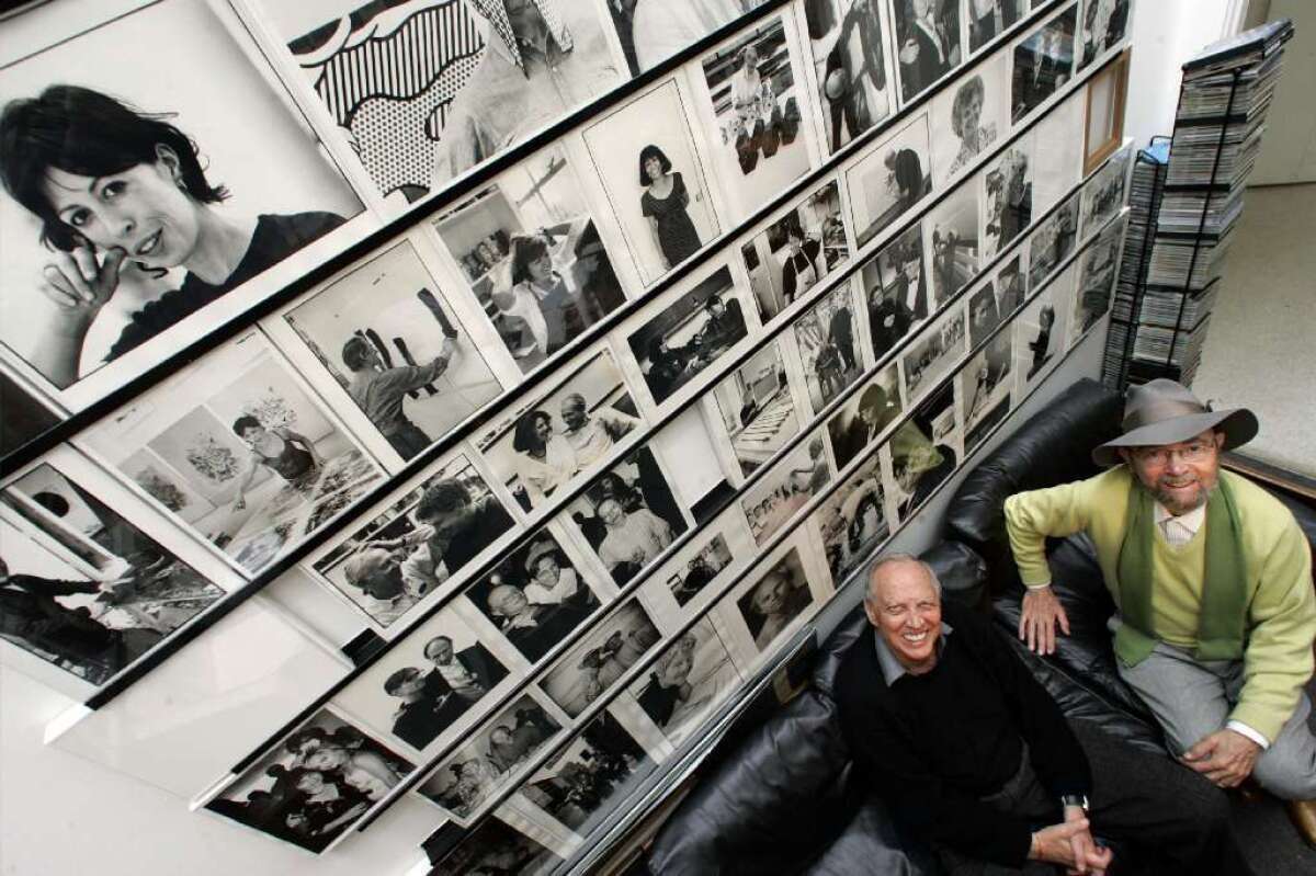 Stanley Grinstein and Sidney Felsen sit on a couch beside a wall of photographs and look up at the photographer.
