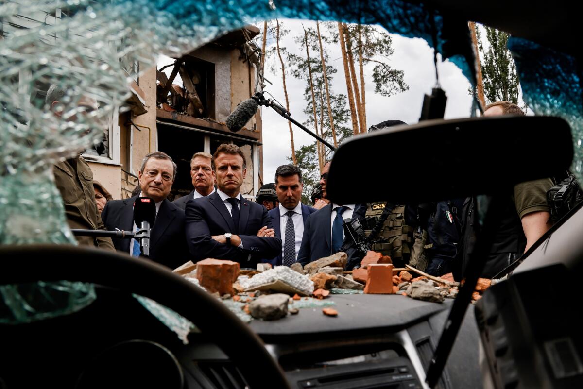 Several men in suits, framed by rubble and a windshield's shattered glass, seen from the driver's seat of a damaged vehicle.