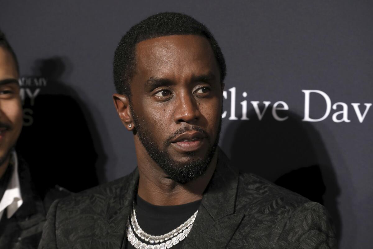 Sean Combs wearing a black jacket and shirt and chains around his neck stands in front of a backdrop 