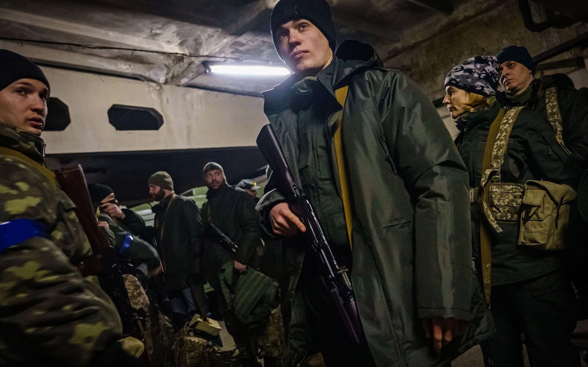 A man in a black beanie and green coat holding a rifle, surrounded by other armed men and women.