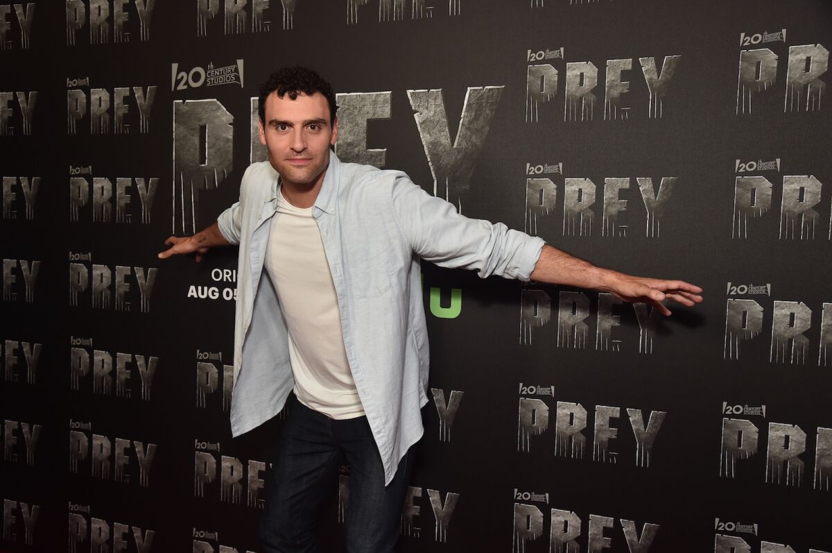 A man spreads his arms while standing in front of a promotional wall for the movie "Prey."