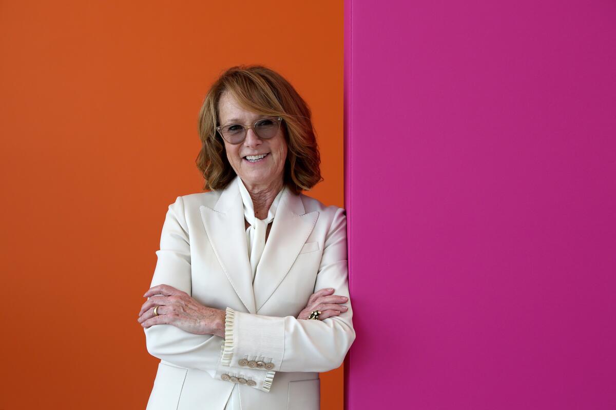 Ann Philbin, in a white suit, leans against a bright fuchsia wall, a bright orange wall behind her.