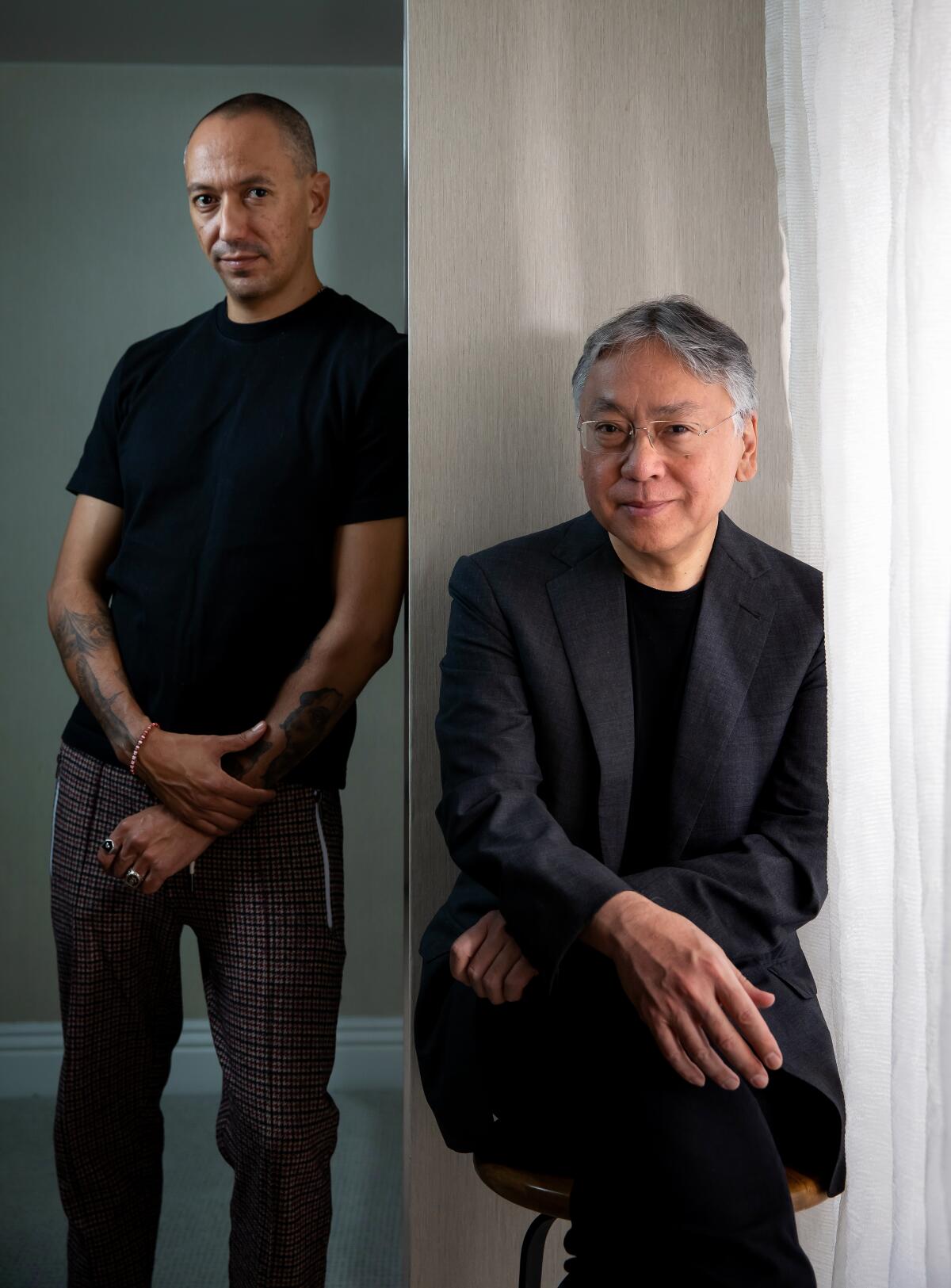 A man sits on a chair in front of a partial wall while a second man leans against the wall for a portrait.