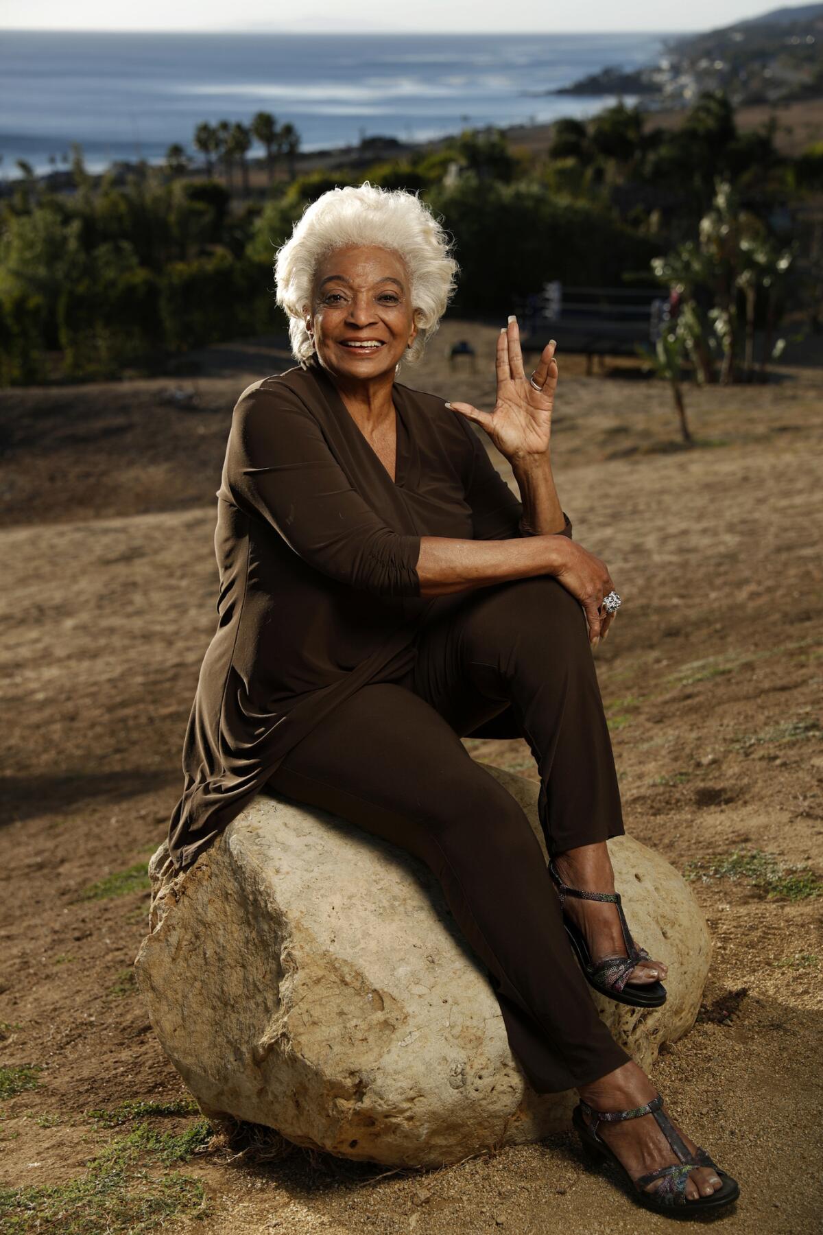 A woman with white hair sits on a rock and flashes the Vulcan salute.