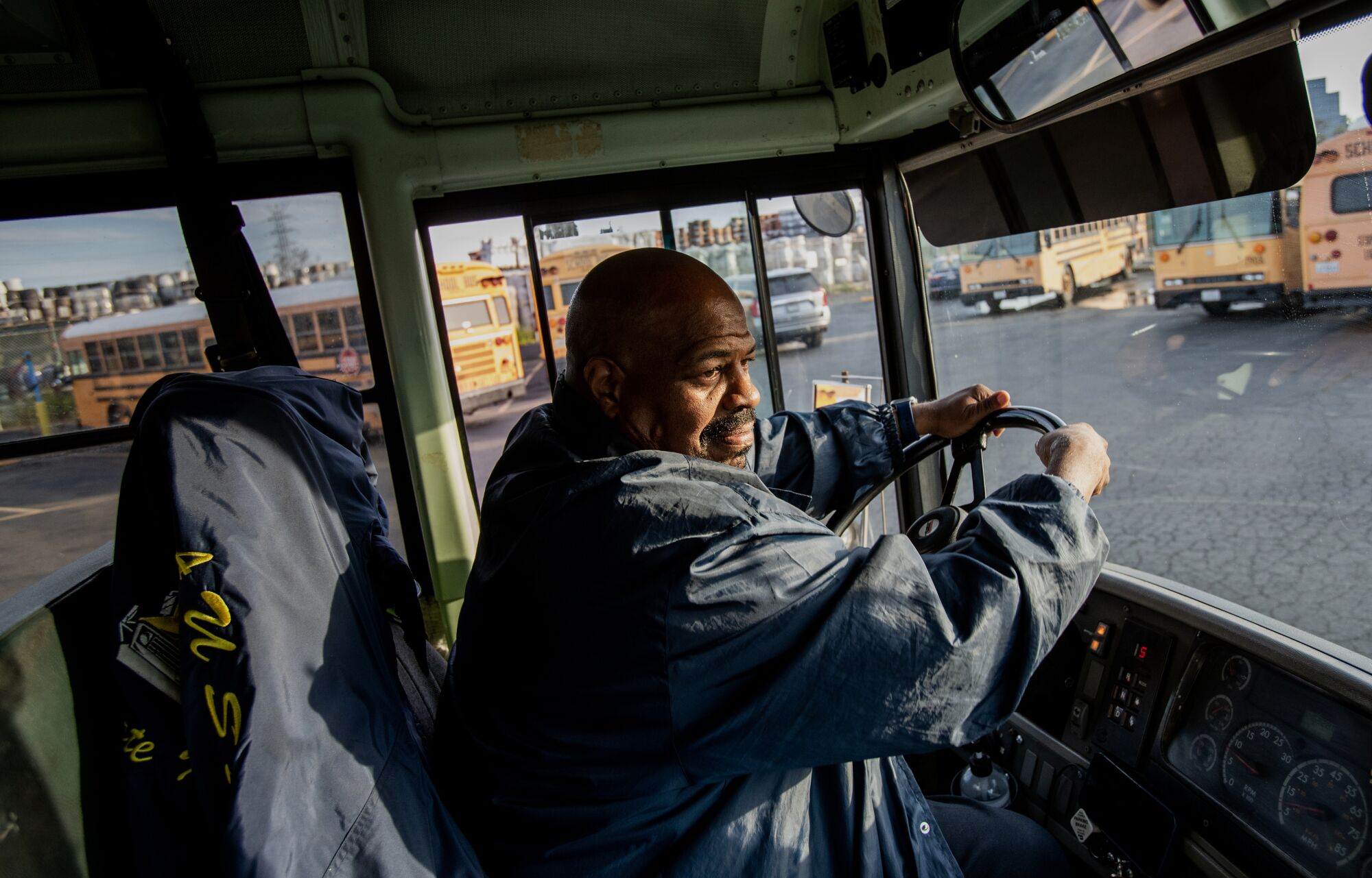 A man sits in the driver's seat of a school bus, holding onto the steering wheel, partially lighted by sun.