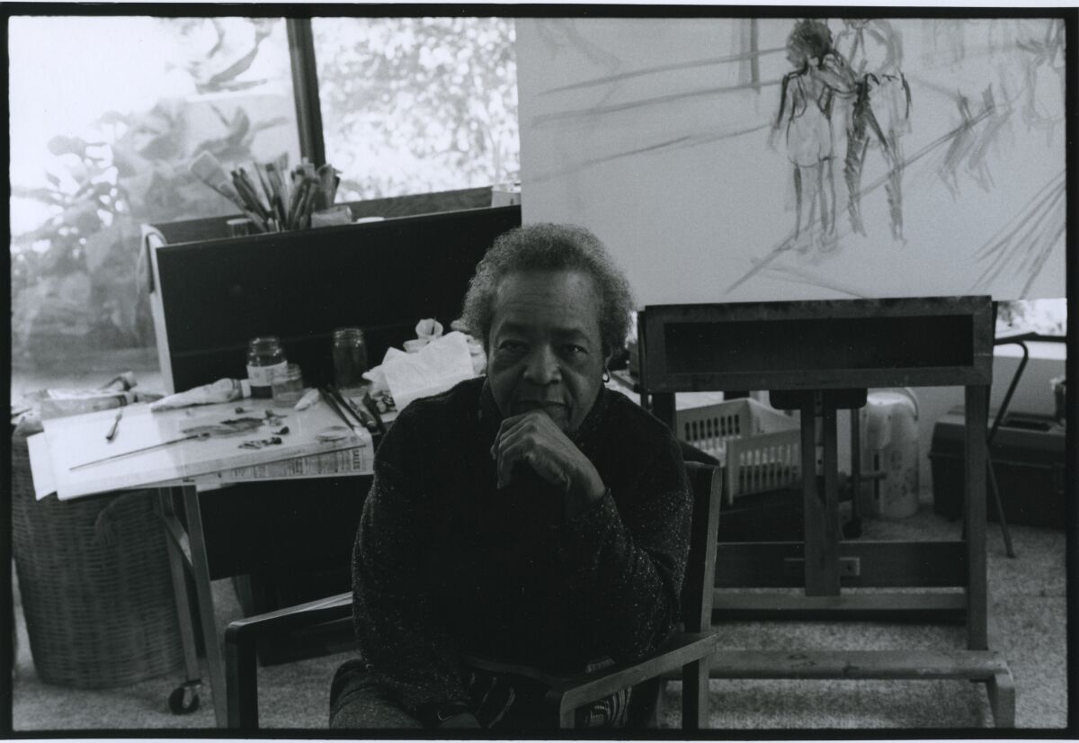 A black and white image shows Samella Lewis sitting before a studio table, an incomplete drawing behind her