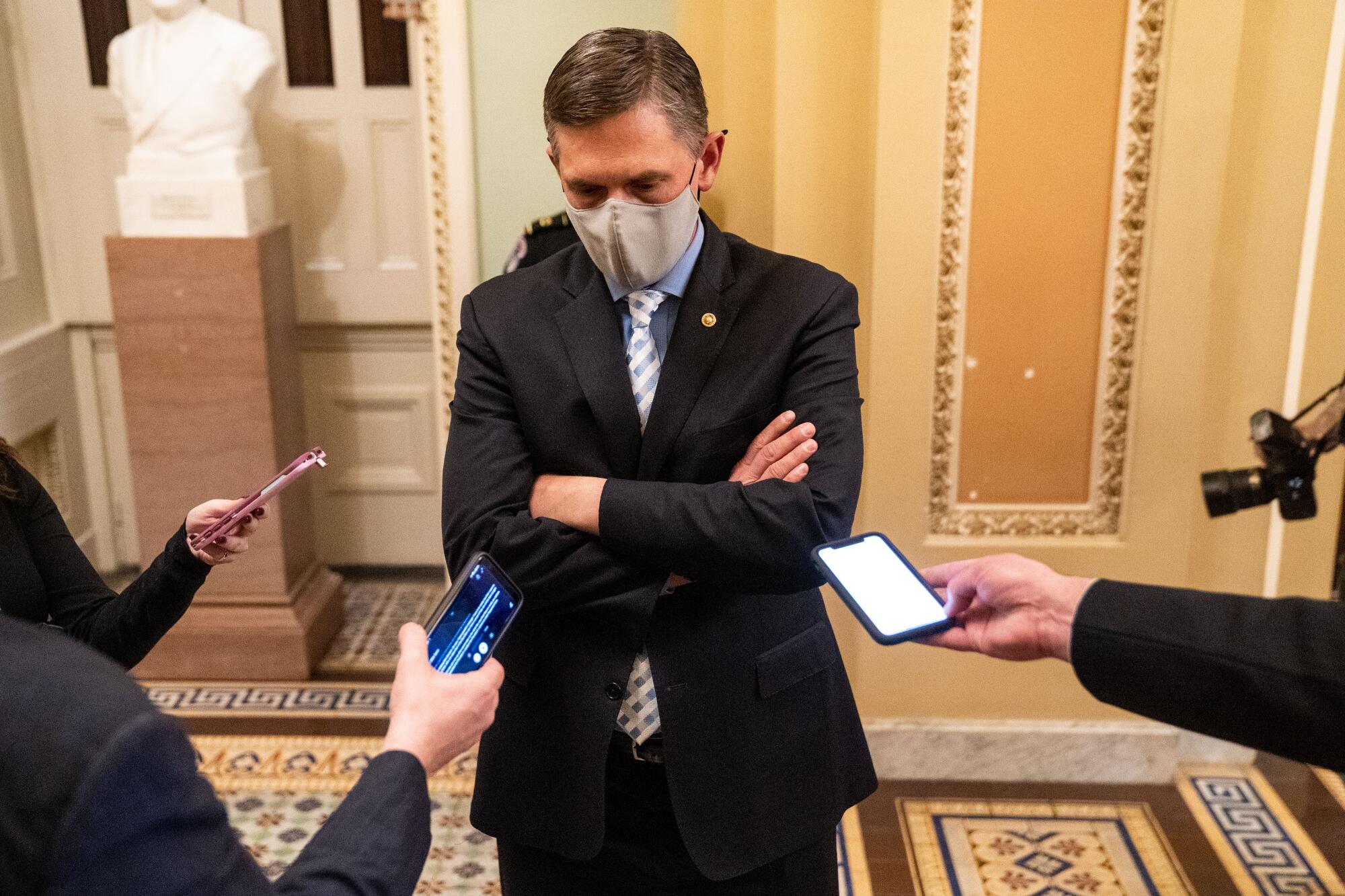 A senator with his arms crossed stands in front of three reporters holding phones out toward him