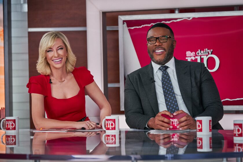 A woman and a man on a TV set, seated at a desk that holds several coffee mugs.