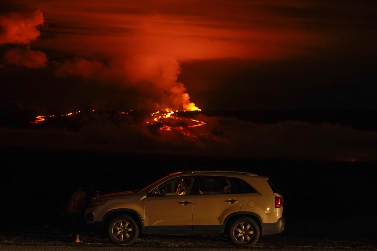 A man talks on a phone in his car at night with lava, flames and smoke in the distance.