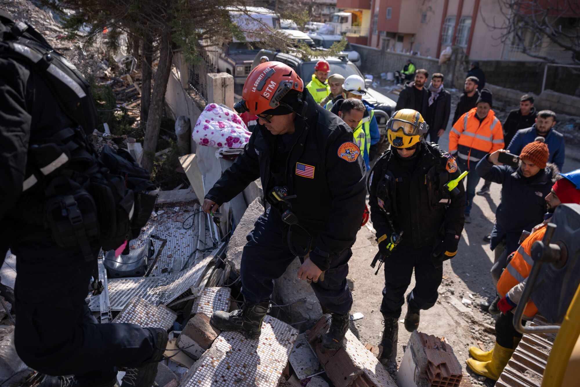 Des personnes portant des casques enjambent les décombres au milieu des bâtiments pendant la journée.