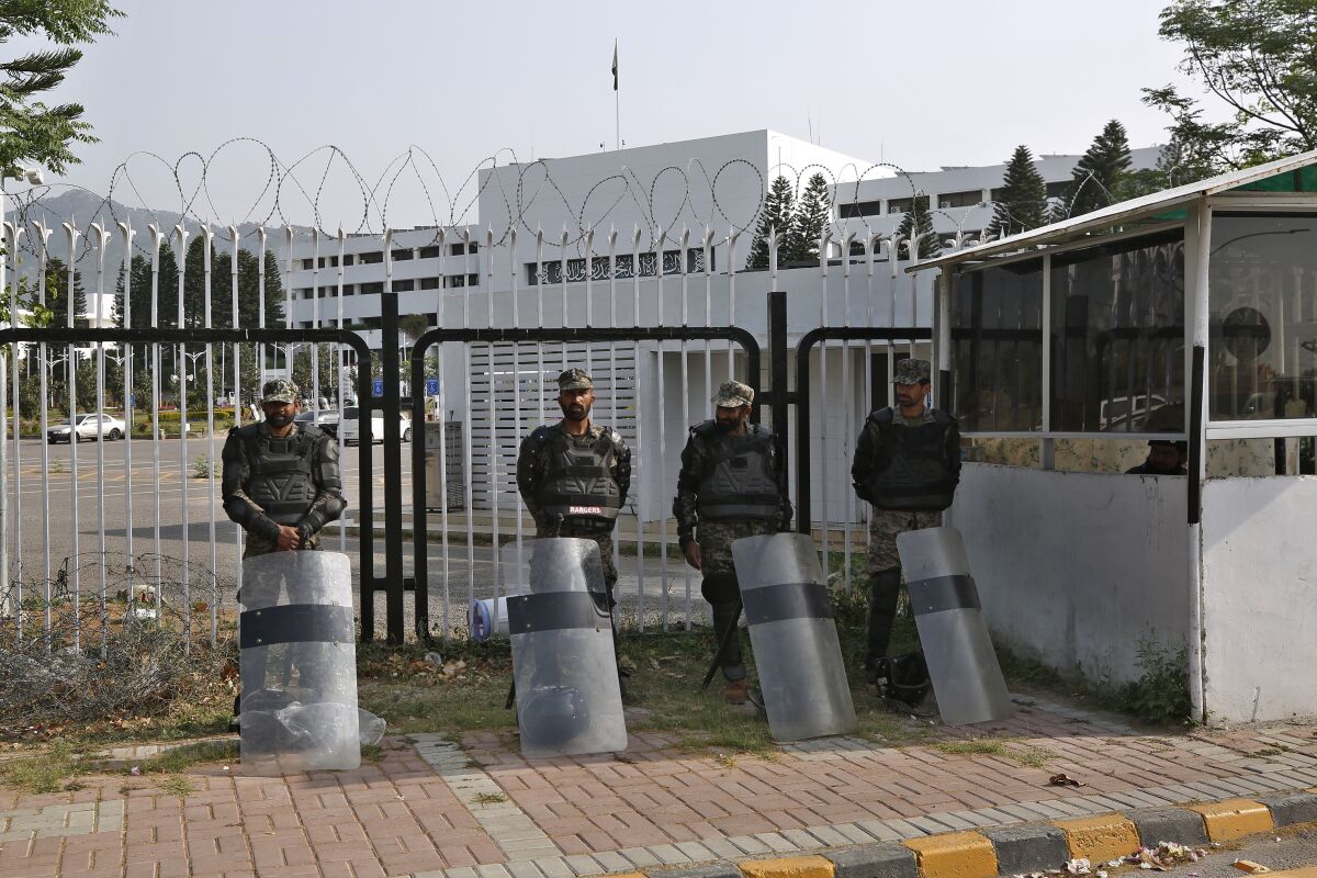 Pakistani paramilitary troops stand guard with riot gear outside gates with barbed wire atop them.