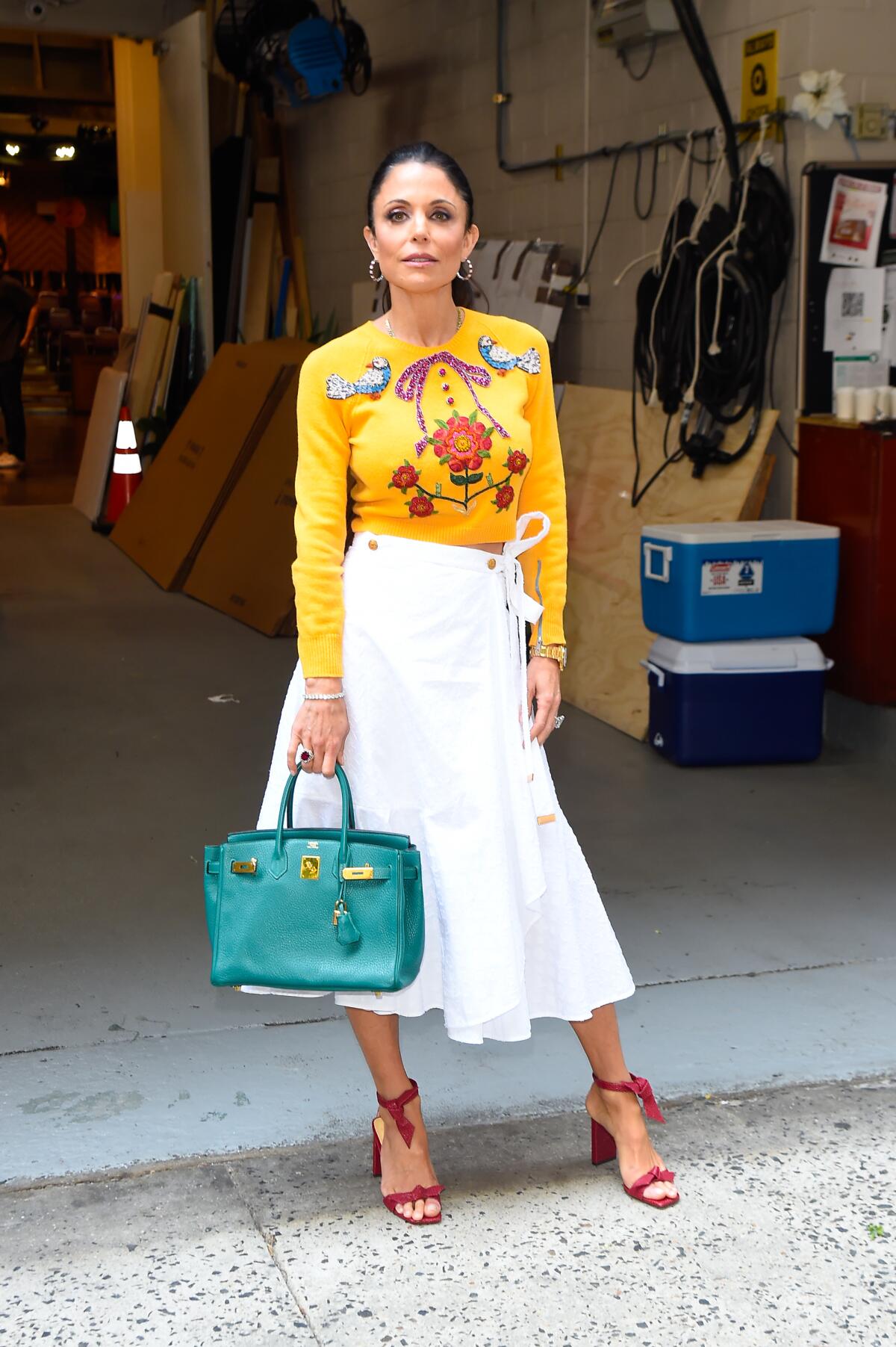 Bethenny Frankel holds a teal handbag and is wearing a yellow sweater and long white skirt.