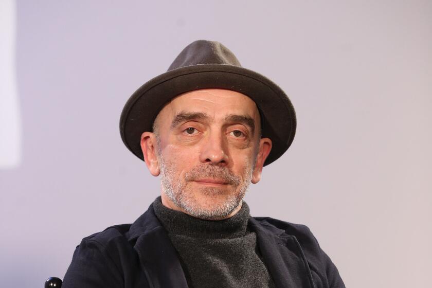 A man with short, gray facial hair in a wide-brimmed hat, a dark sweater and shirt sitting with his hands on his lap 