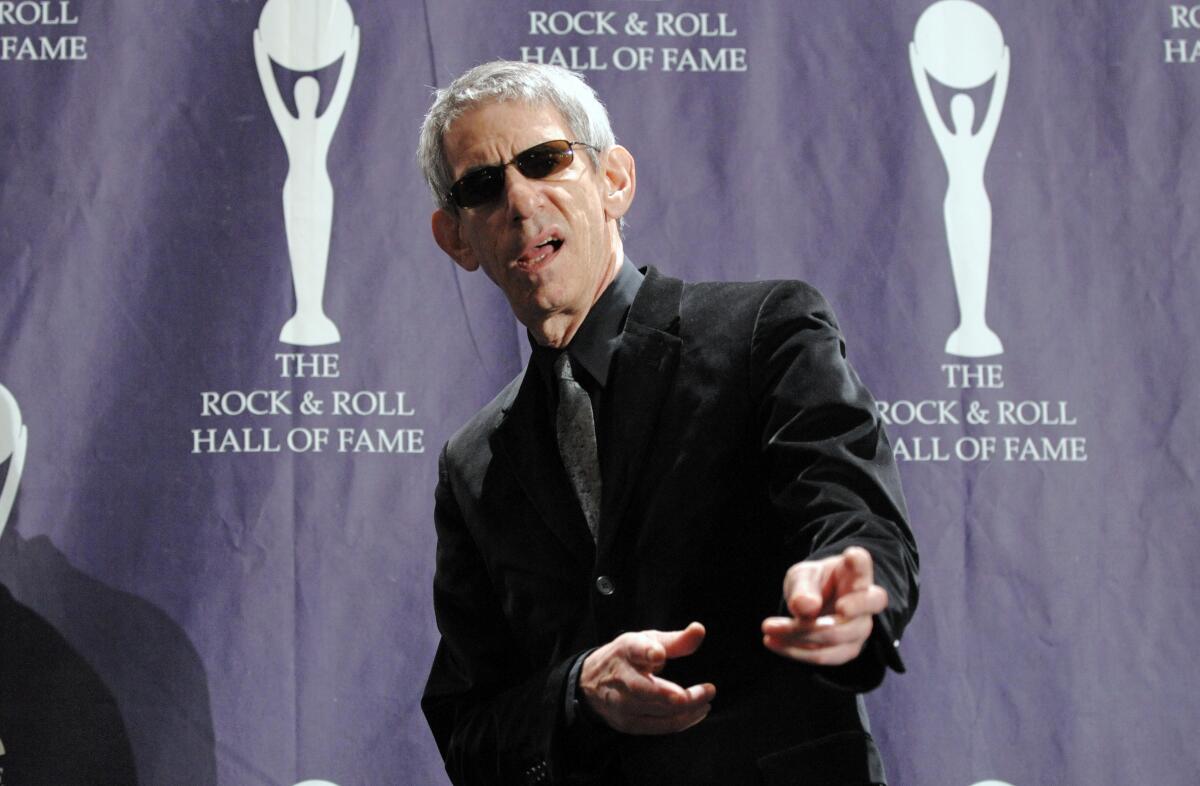 A man with gray hair wearing sunglasses and a black suit points finger guns toward the camera