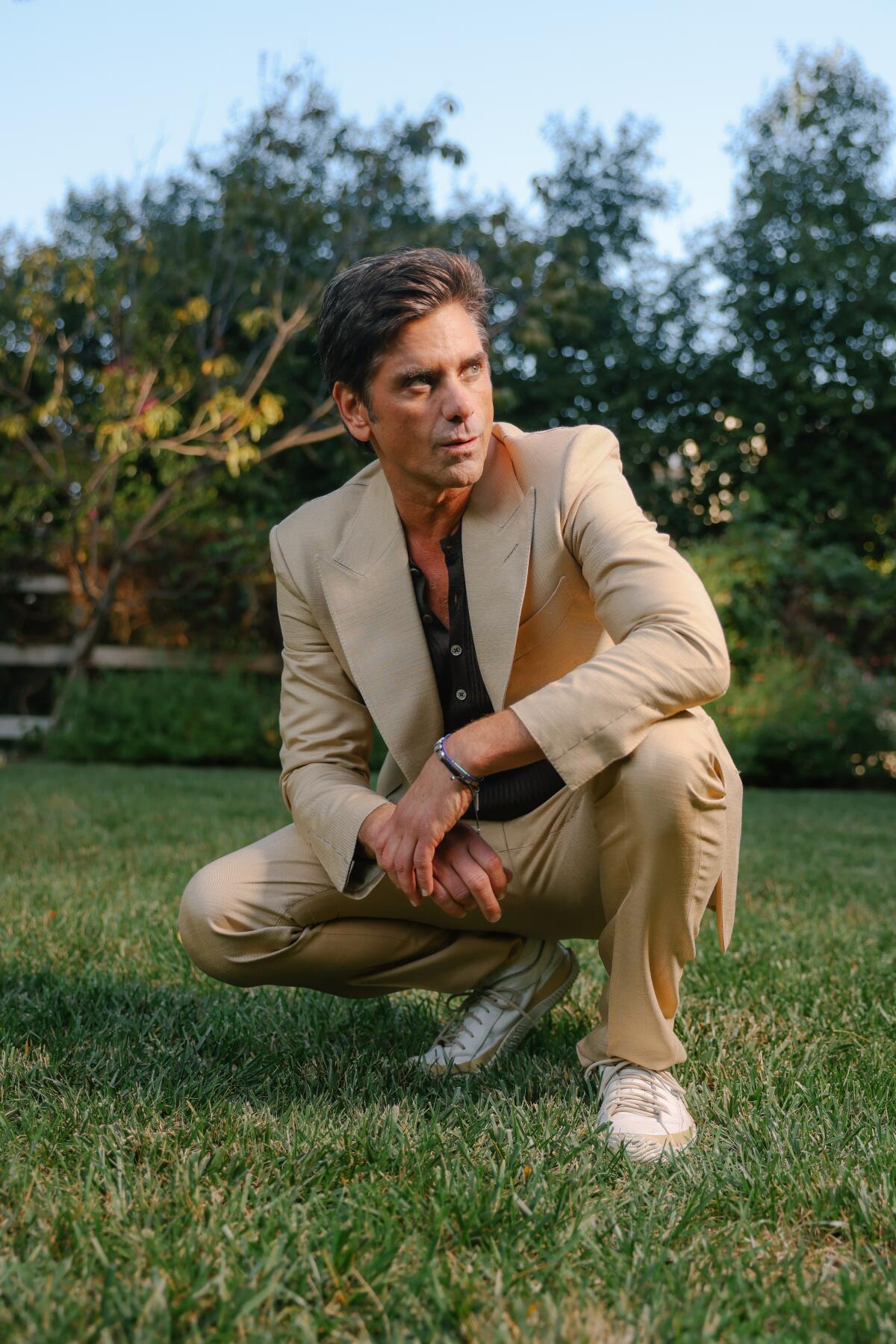 John Stamos, in a tan suit and white sneakers, squats in a green yard with tree in the background.