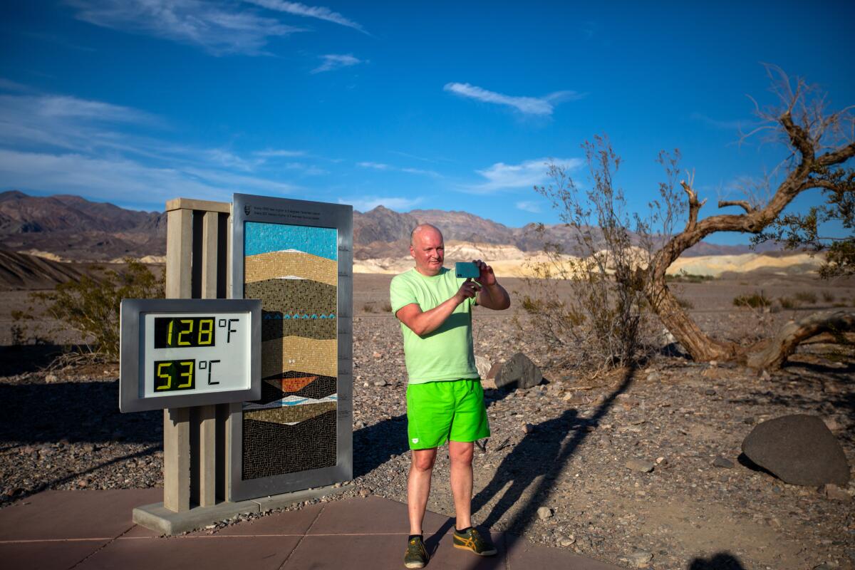 A man wearing bright green shorts and T-shirt takes a selfie in front of a digital thermometer that says128 degrees.