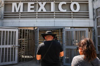 A man in a dark shirt and hat and a woman with brown hair walk toward turnstiles under a sign that reads MEXICO