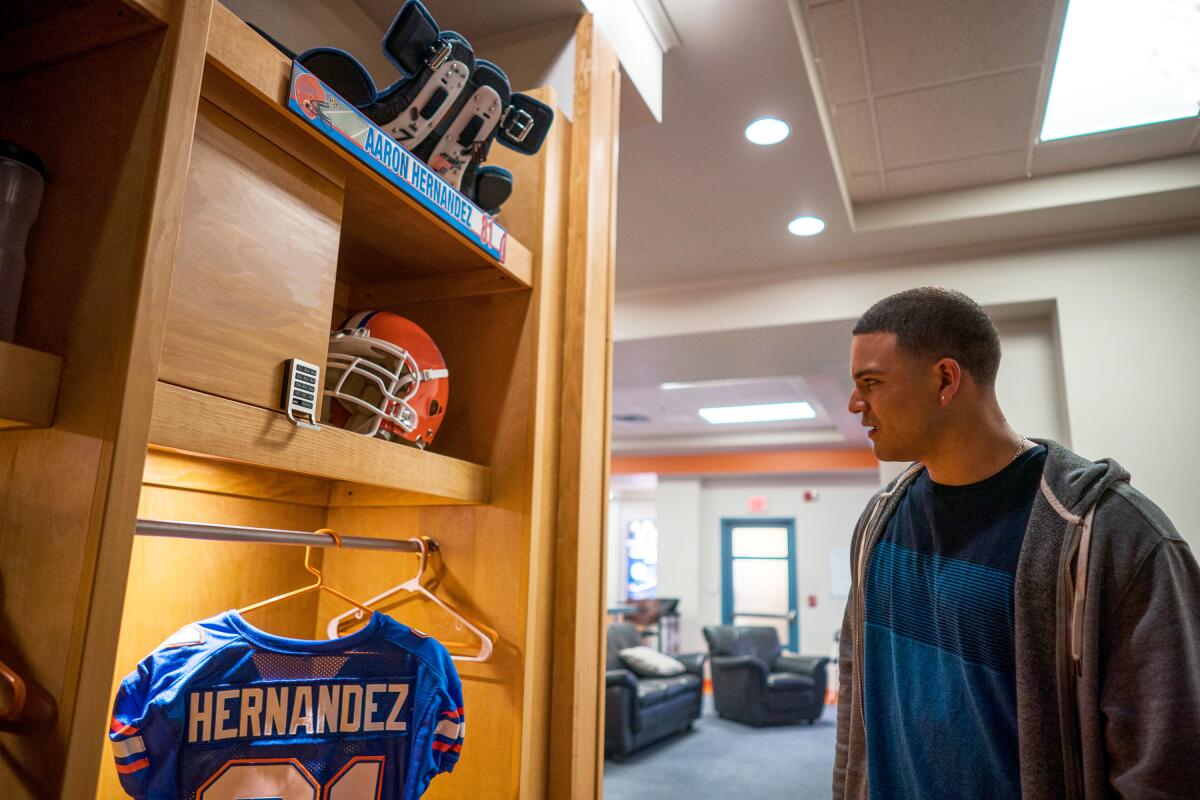 A man in a jacket looks at a football jersey with the name Hernandez on it hanging in a locker room stall.