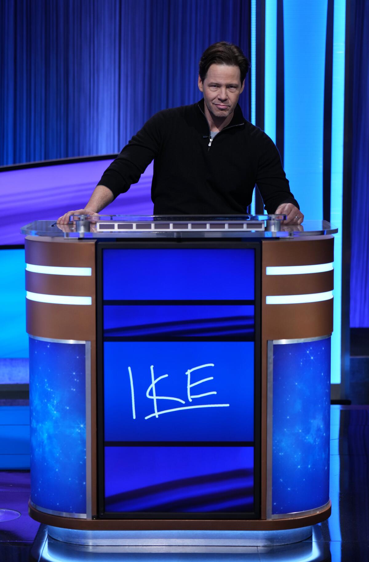 Ike Barinholtz stands at a lectern on a game show with his name on the front