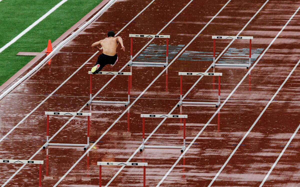 A shirtless man jumps hurdles on a track in the rain.