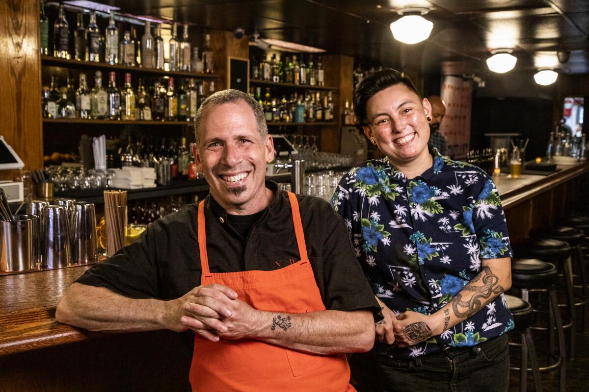 Two smiling people lean against a bar in a restaurant.