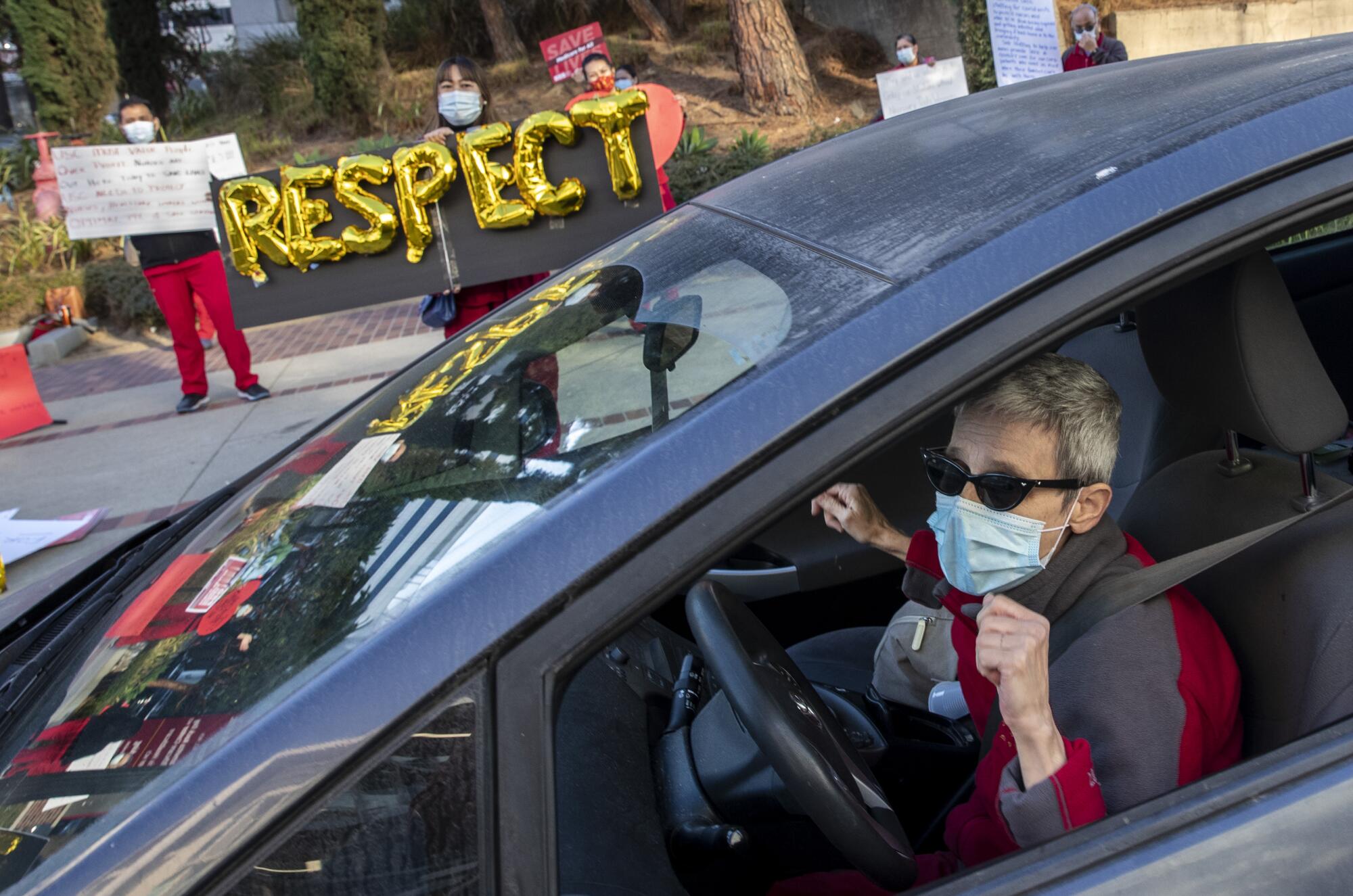 Protesters wearing face masks stand near a sign reading "Respect" as a woman sits in the driver's seat of a car.