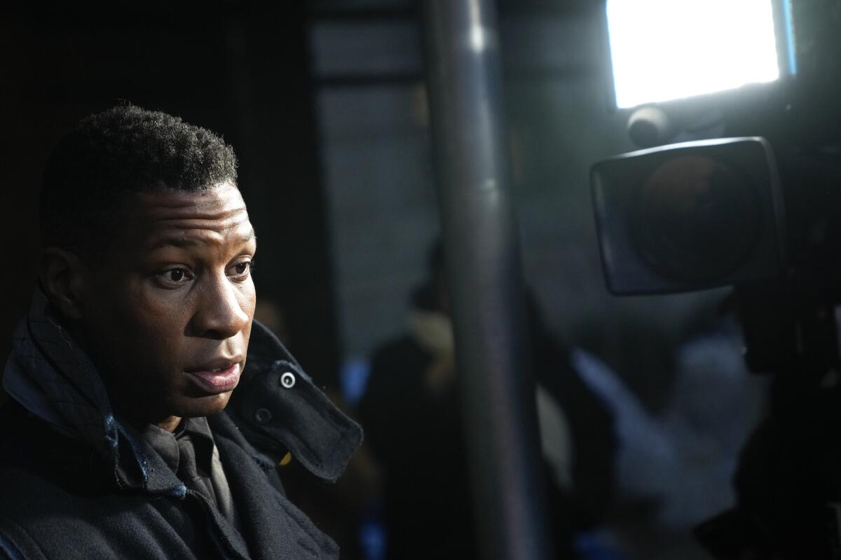 Jonathan Majors looks stunned as he walks out of a courthouse in a black coat and passes a media camera