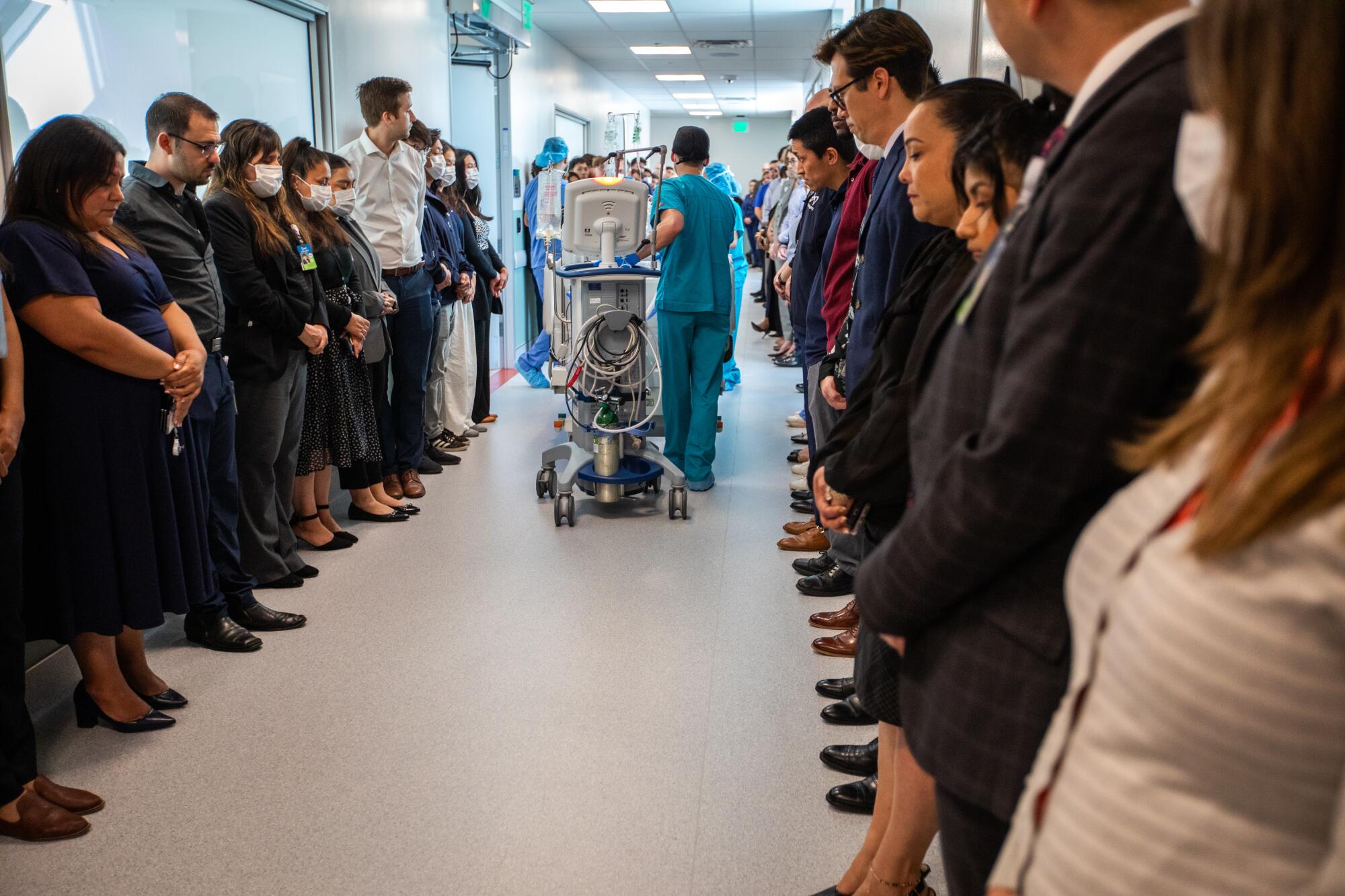 People line a hallway as staff in scrubs push through with a hospital bed flanked by a stand with a monitor.