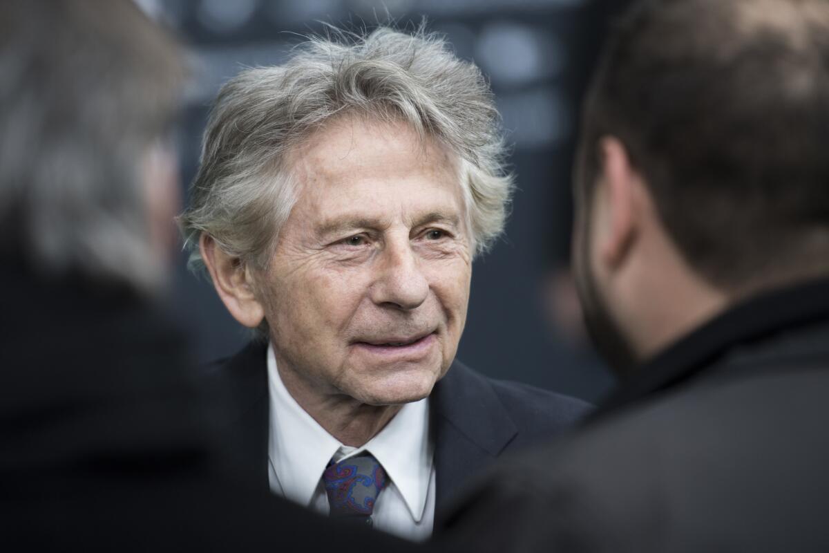 Roman Polanski wearing a black suit with a white shirt and blue tie