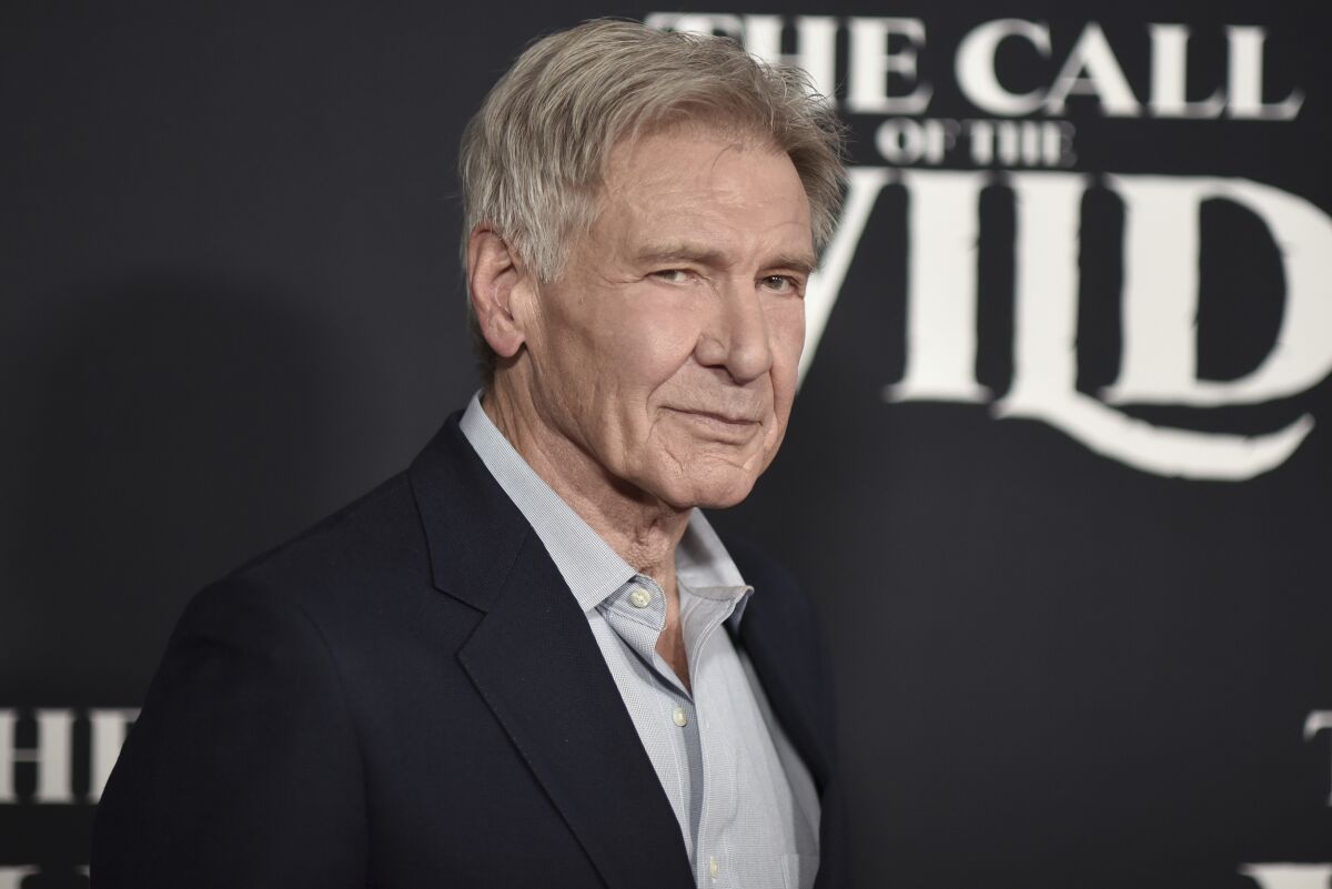 An older man in a gray shirt and black blazer smirks and gives side-eye at a premiere