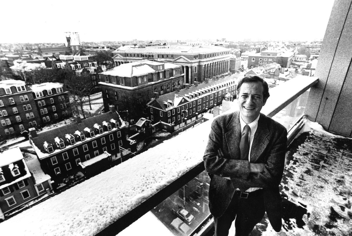 A black-and-white photo of a man in a suit standing on a balcony in snow, with Harvard University below him.