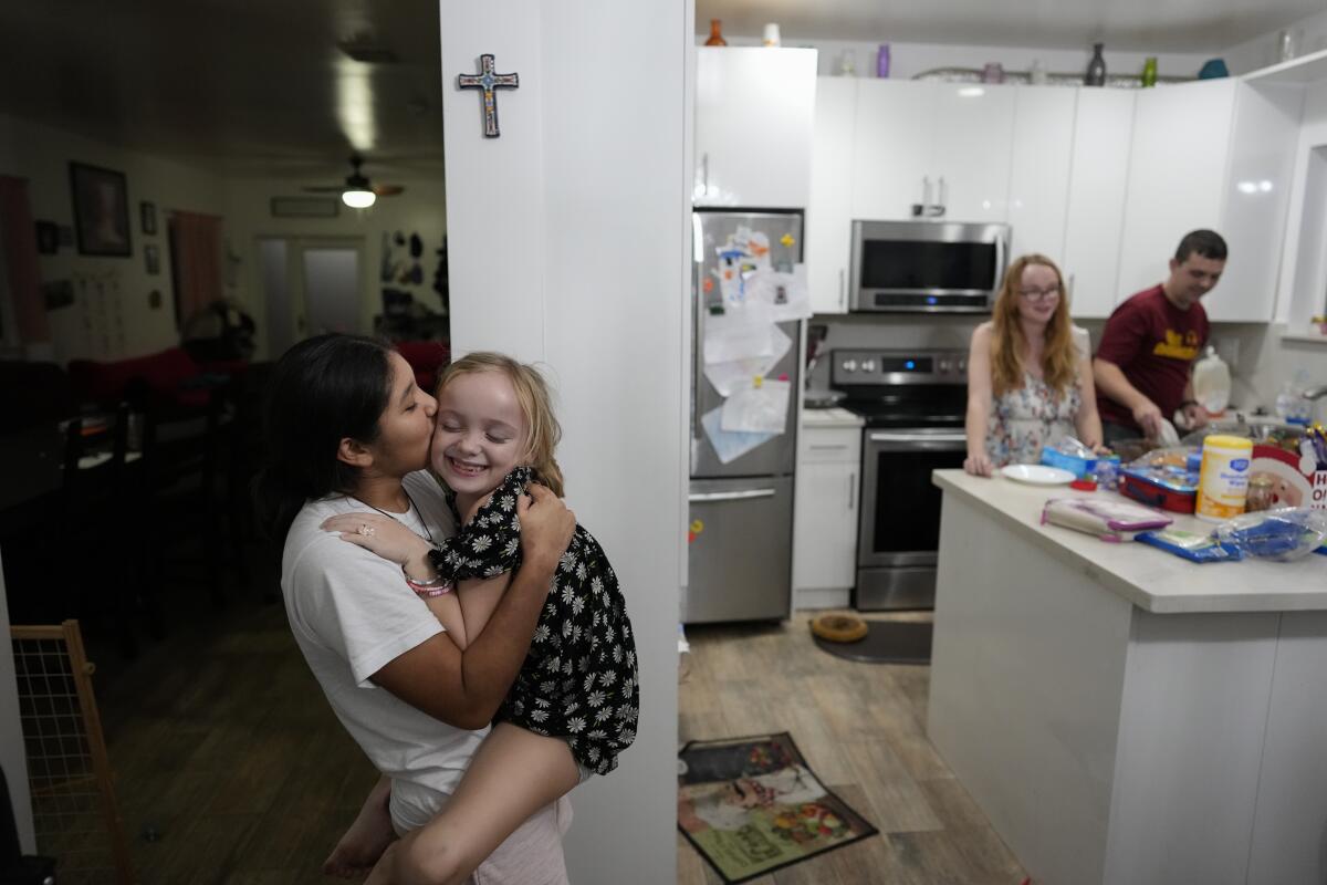 A child kisses a younger child she's holding in her arms as two adults stand behind her in a kitchen.