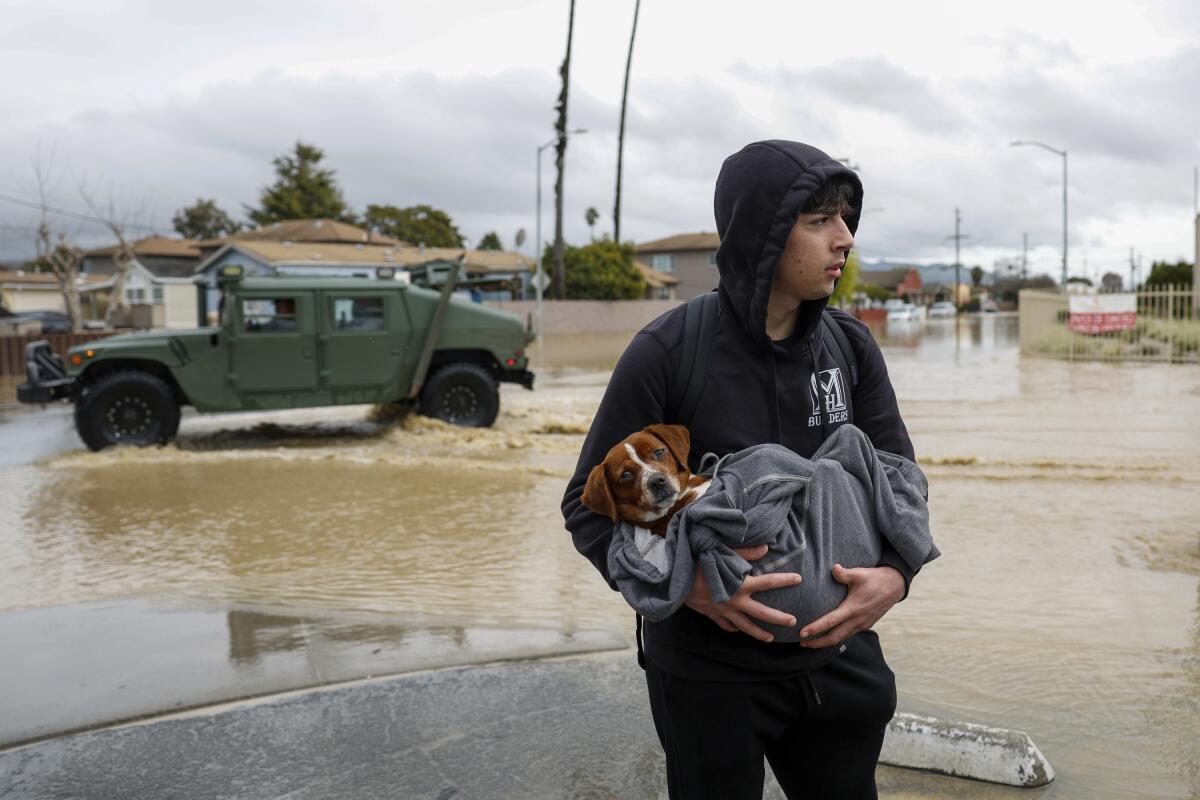 A man holds a dog wrapped in a blanket as a military vehicle drives through flooded streets behind them.