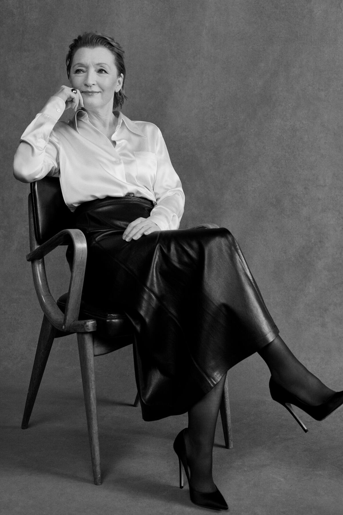 Lesley Manville in a white shirt and dark skirt sits on a chair with her hand resting on the side of her face.