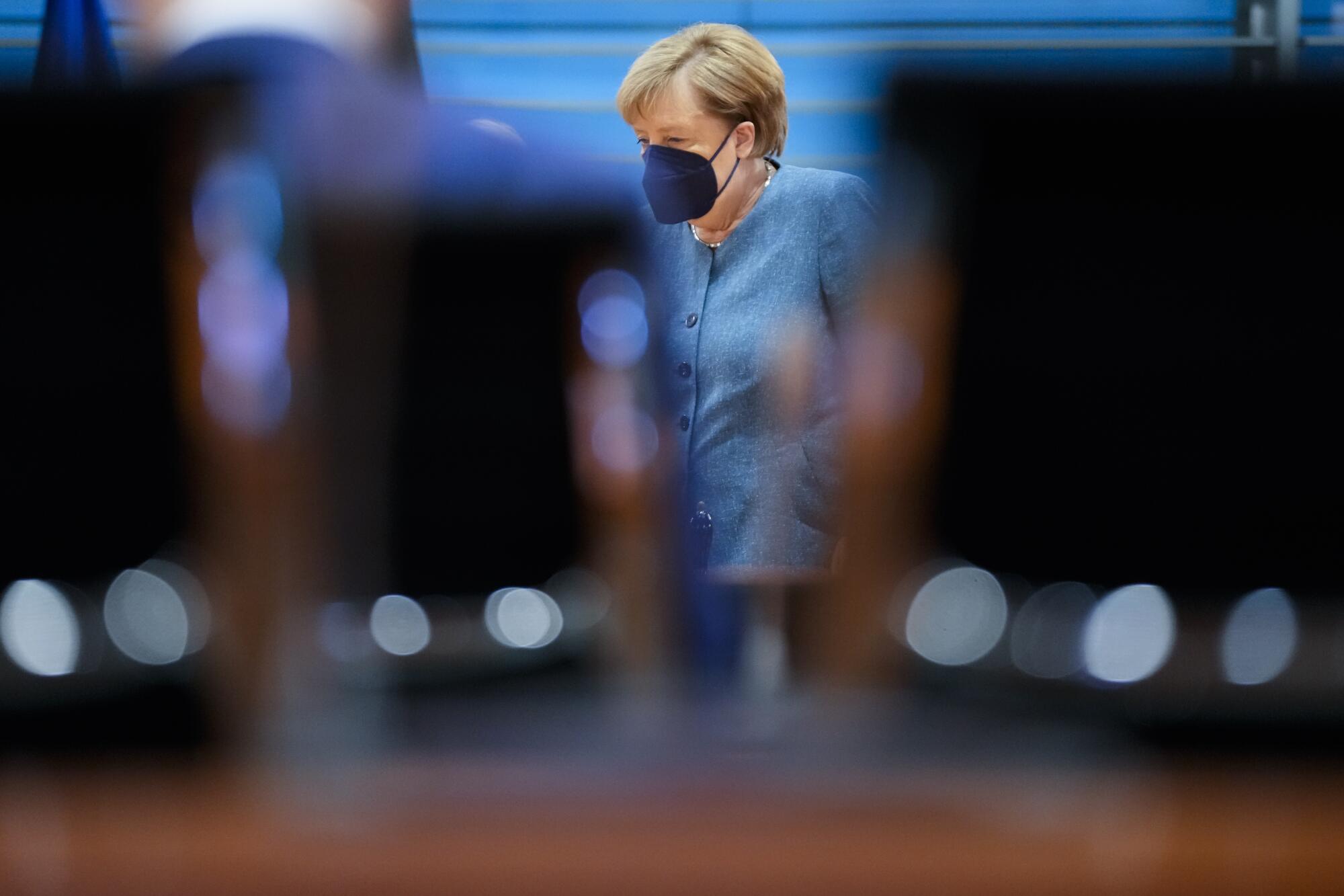Angela Merkel, wearing a light blue jacket and dark mask, is seen through a gap between a man and a chair
