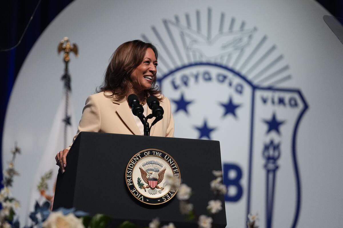 Kamala Harris speaking from a lectern with the vice presidential seal, a large white and blue sorority logo in the background
